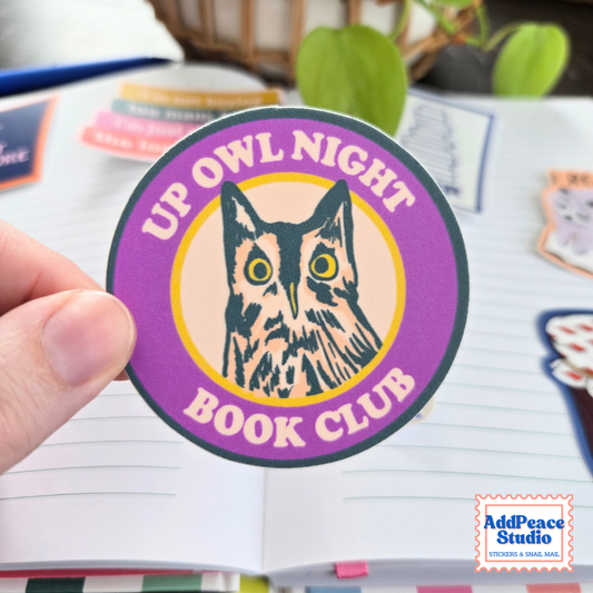 A hand holds a round sticker that reads 'UP OWL NIGHT BOOK CLUB' with an owl illustration in the center. The sticker has a purple outer ring, yellow and peach center, and is shown over an open notebook with other stickers in the background.