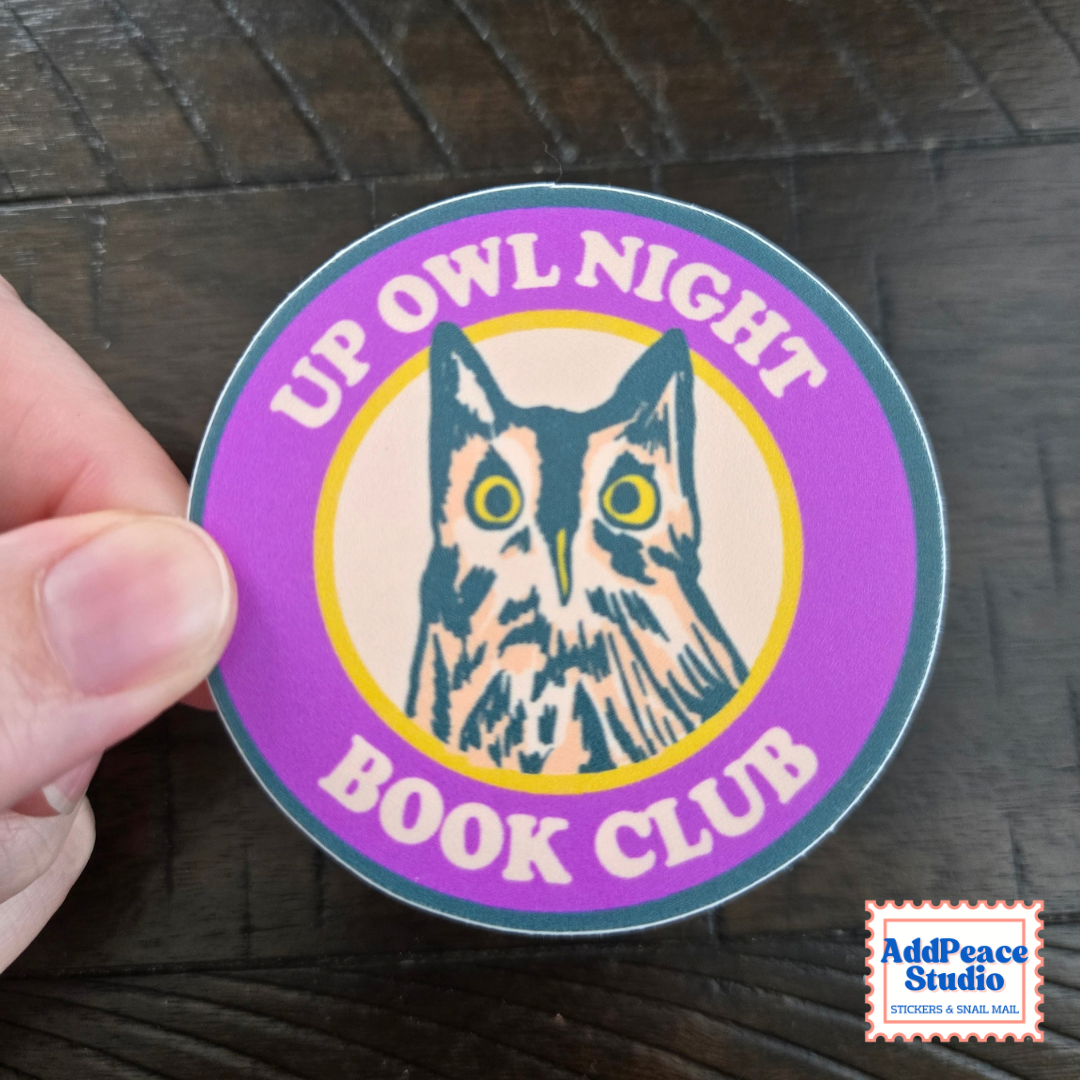 A round sticker held by a hand against a dark wood background. The sticker features a purple outer ring, a beige center, and a dark teal owl illustration with yellow eyes.