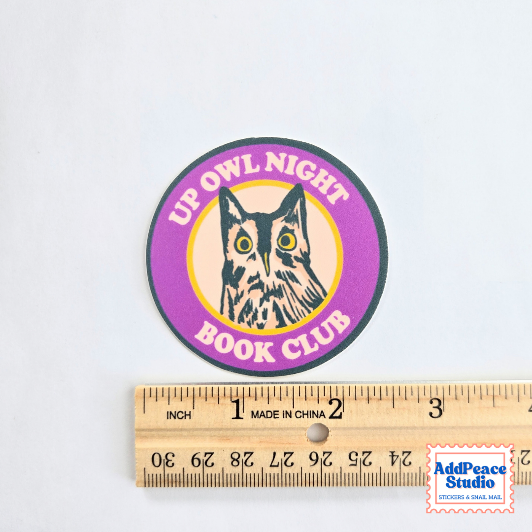 A round sticker reading “UP OWL NIGHT BOOK CLUB” with an owl illustration, shown above a wooden ruler to indicate size. The sticker has a purple outer ring, yellow center, and black text and outline details.