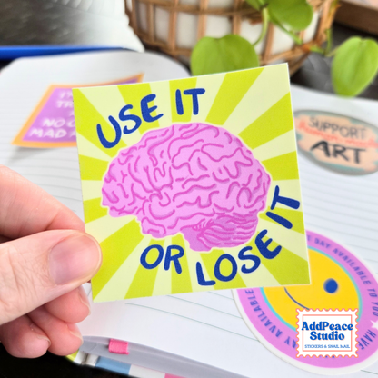 A hand holds a square sticker featuring a pink brain illustration on a lime sunburst background with the text 'USE IT OR LOSE IT' in blue. The sticker is shown over a notebook with other stickers visible in the background.