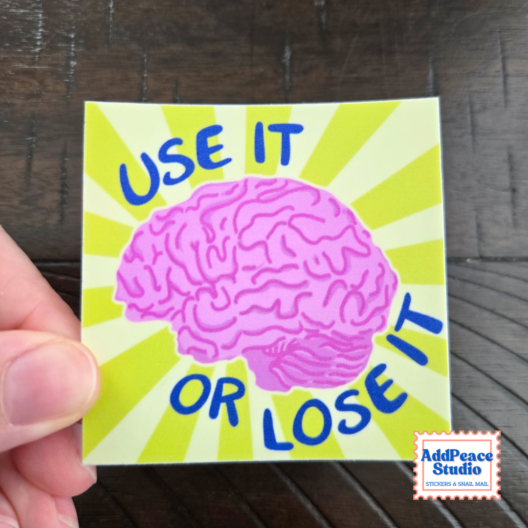 A hand holds a square sticker with a pink brain illustration and the text 'USE IT OR LOSE IT' in blue on a lime green sunburst background, set against a dark wood surface. AddPeace Studio logo is visible in the bottom right corner.