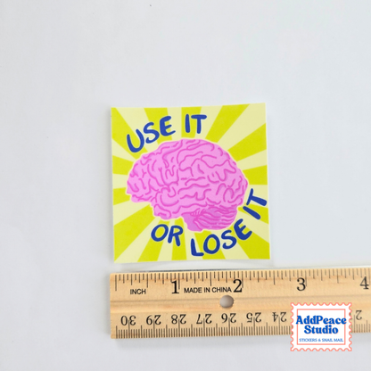 A square sticker featuring a pink brain illustration on a yellow sunburst background with the text 'USE IT OR LOSE IT' in blue. A wooden ruler is placed below the sticker to show scale, and an AddPeace Studio logo watermark appears in the bottom right corner.