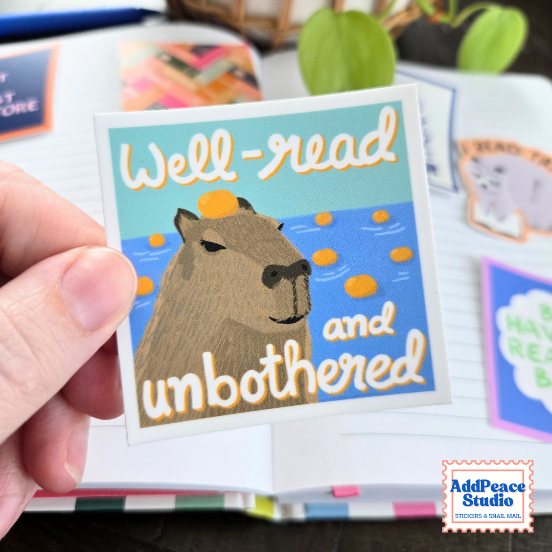 A hand holds a square sticker featuring a capybara in blue water with the text 'Well-read and unbothered'. The sticker is shown in a styled desk setting with notebooks and other stickers in the background.