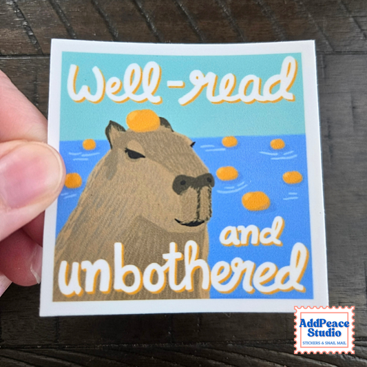 A hand holds a square sticker featuring a capybara illustration with the text 'Well-read and unbothered' on a dark wood background. The sticker has a white border and a small 'AddPeace Studio' logo in the bottom right corner.