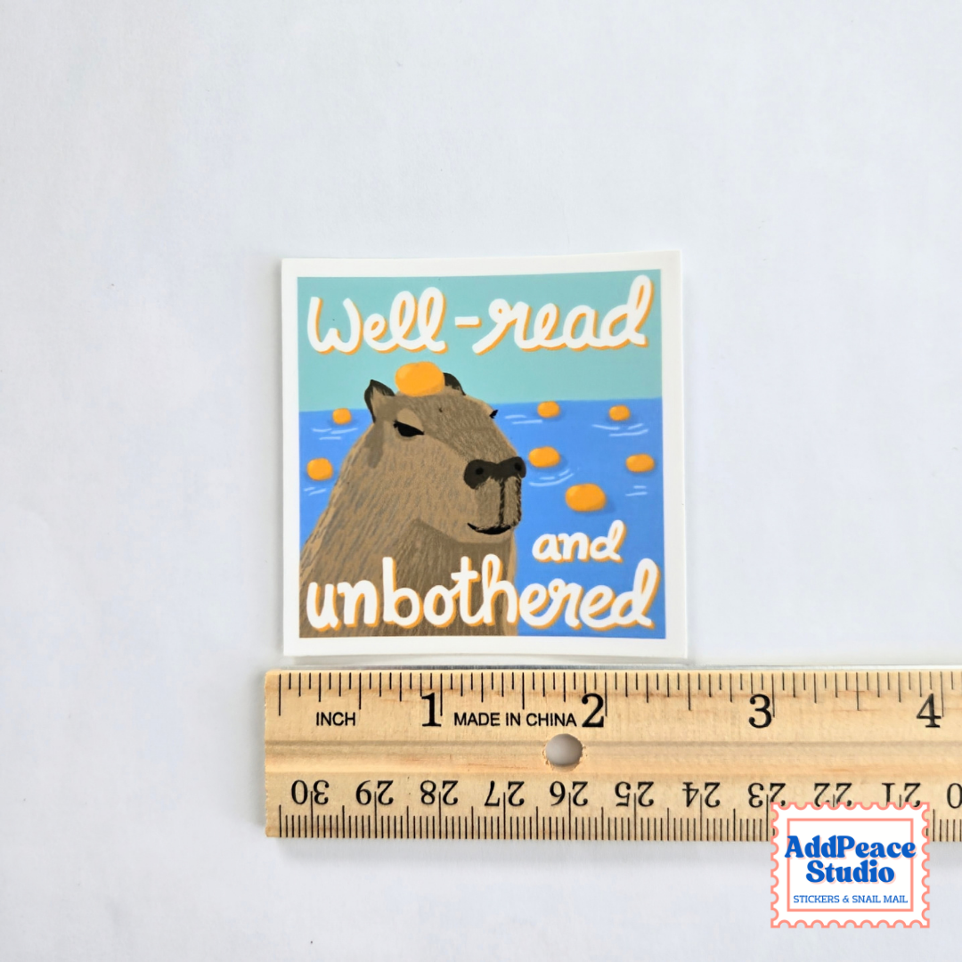 A square sticker with a cartoon capybara and the text 'Well-read and unbothered' placed above a wooden ruler to show its size. The sticker has a white border and is photographed on a light gray background.