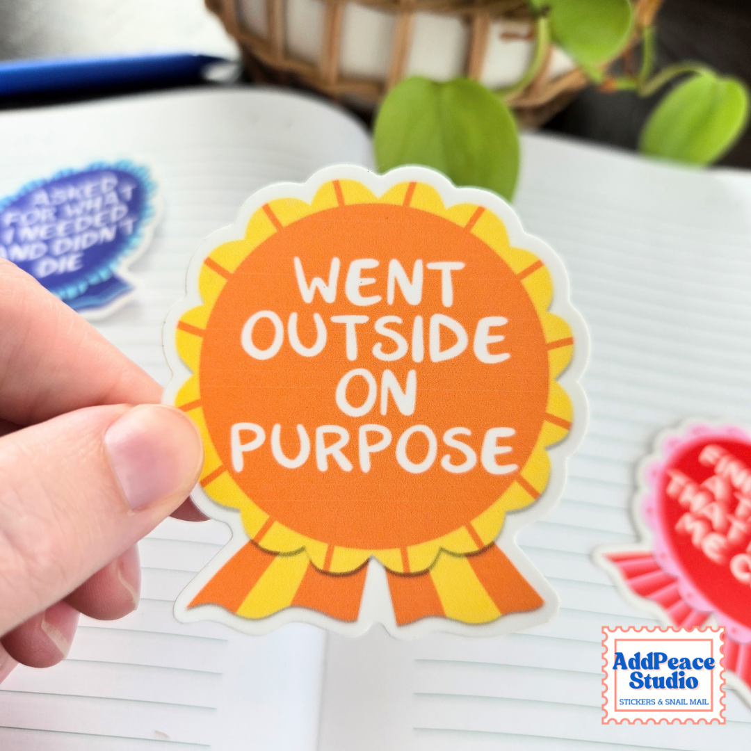 A hand holding an orange award ribbon die cut sticker with white text that says Went outside on purpose