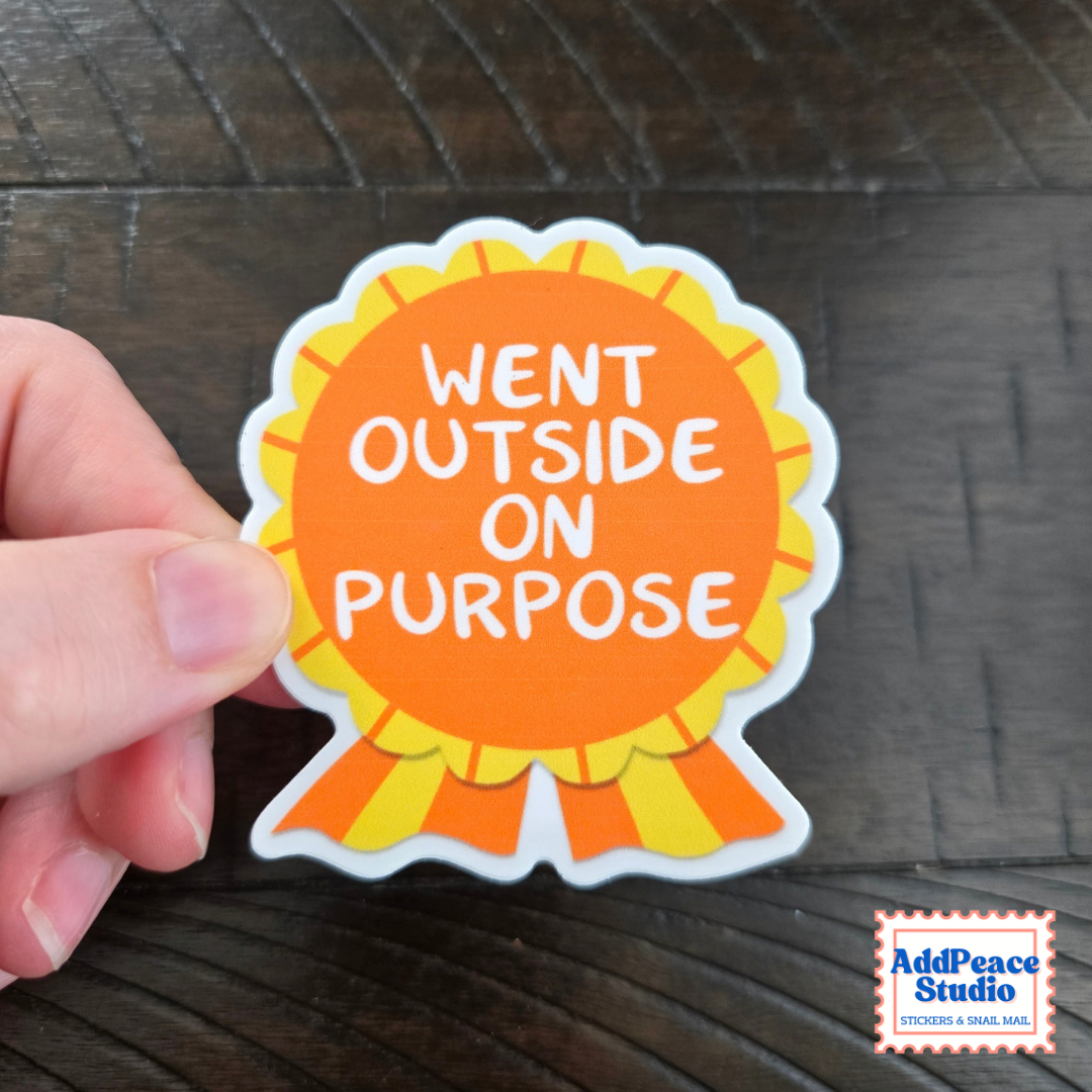 A hand holds a die-cut sticker shaped like a yellow and orange ribbon with the text 'WENT OUTSIDE ON PURPOSE' in white lettering, set against a dark wood background. An 'AddPeace Studio' logo is visible in the bottom right corner.