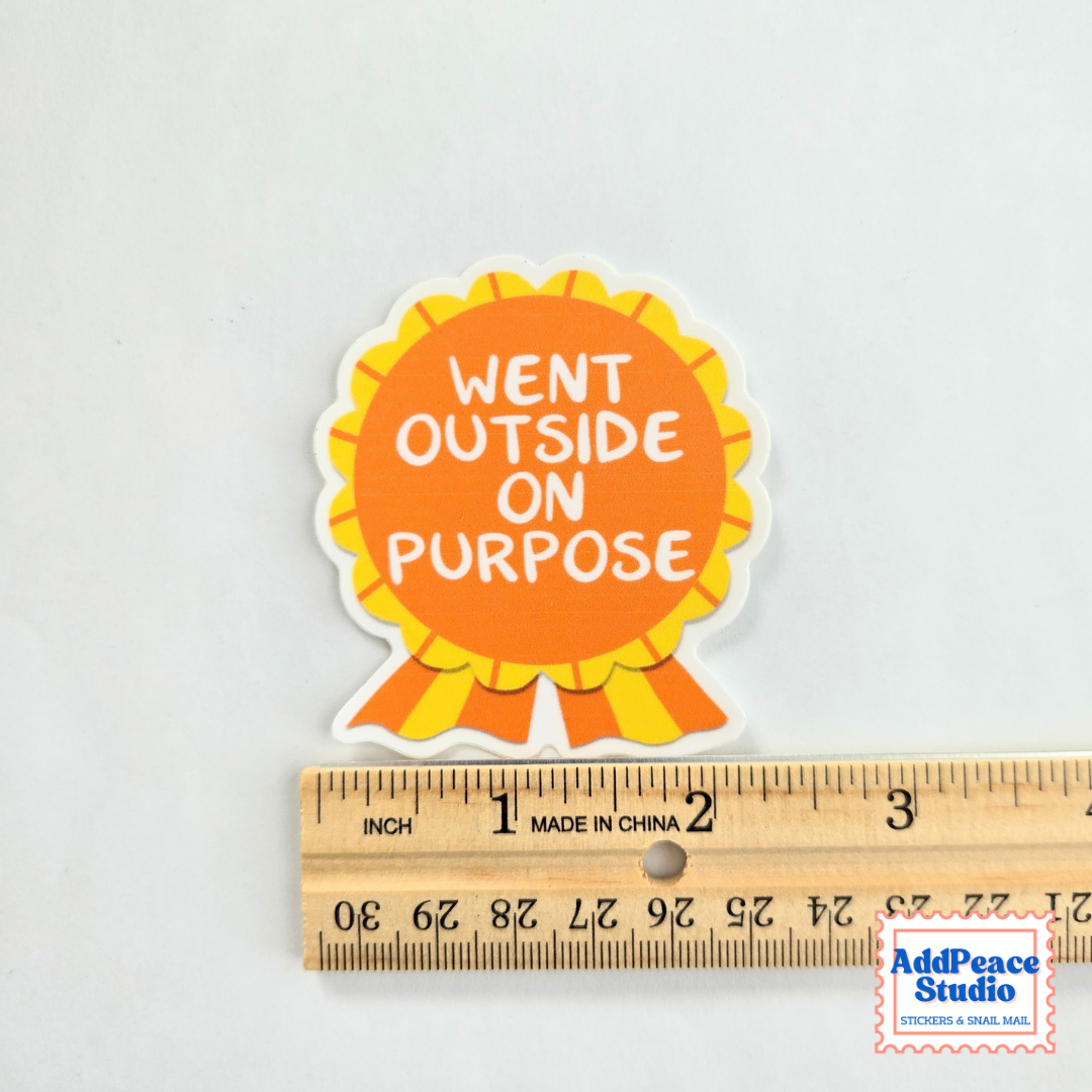 A die-cut sticker with a yellow and orange ribbon design and the text 'WENT OUTSIDE ON PURPOSE' in white, shown above a wooden ruler for scale on a light background.