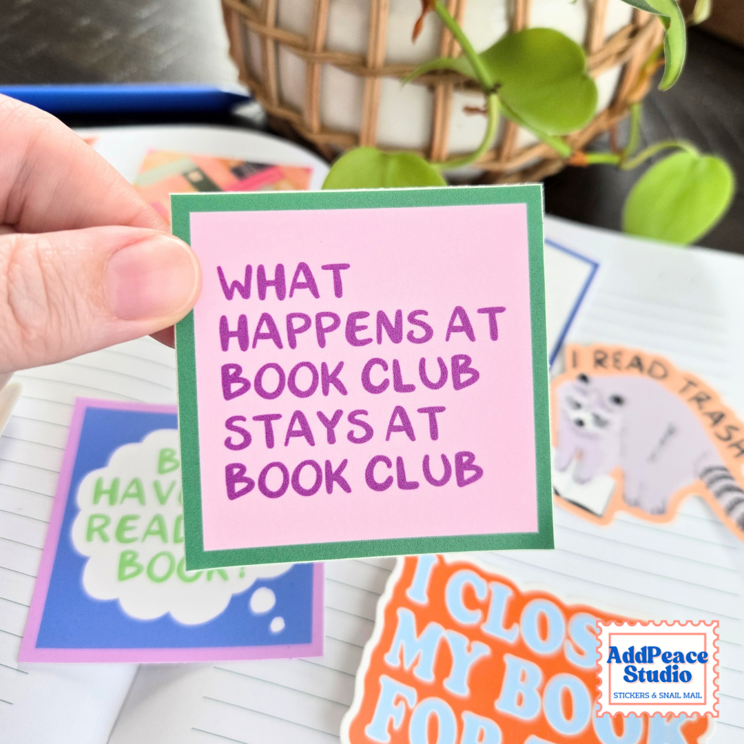 A hand holds a pink sticker with a green border that reads 'WHAT HAPPENS AT BOOK CLUB STAYS AT BOOK CLUB' in purple lettering. Other colorful stickers and a lined notebook are visible in the background, along with a plant and basket.