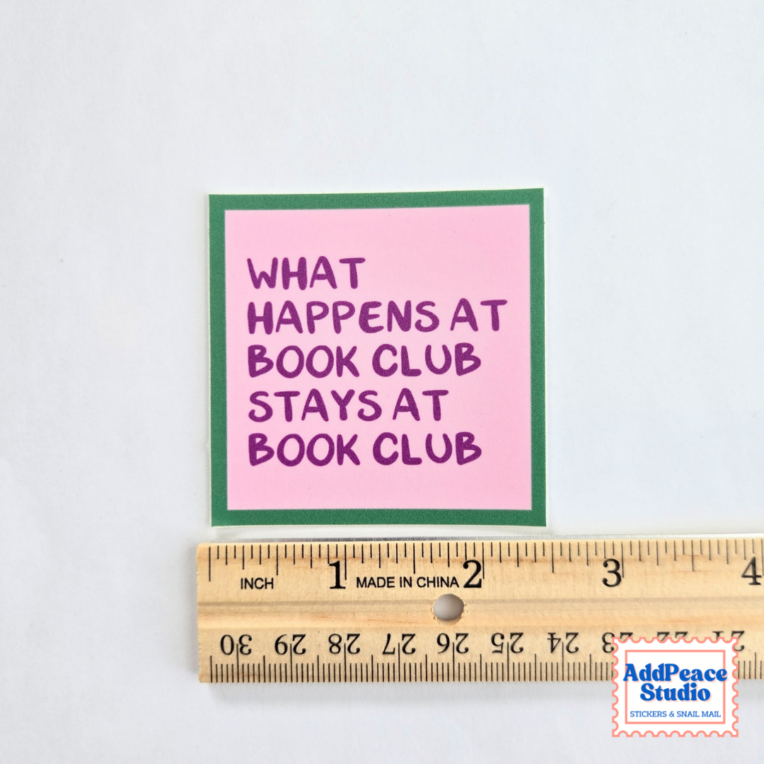 A square sticker with a pink background, green border, and purple text reading 'WHAT HAPPENS AT BOOK CLUB STAYS AT BOOK CLUB' is shown next to a wooden ruler for scale on a white background.