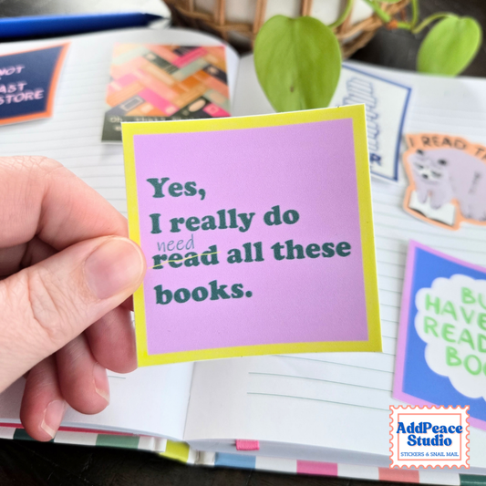 A hand holds a pink square sticker with a yellow border and dark green text that says "Yes, I really do read all these books." Read is marked out and "need" is written in.