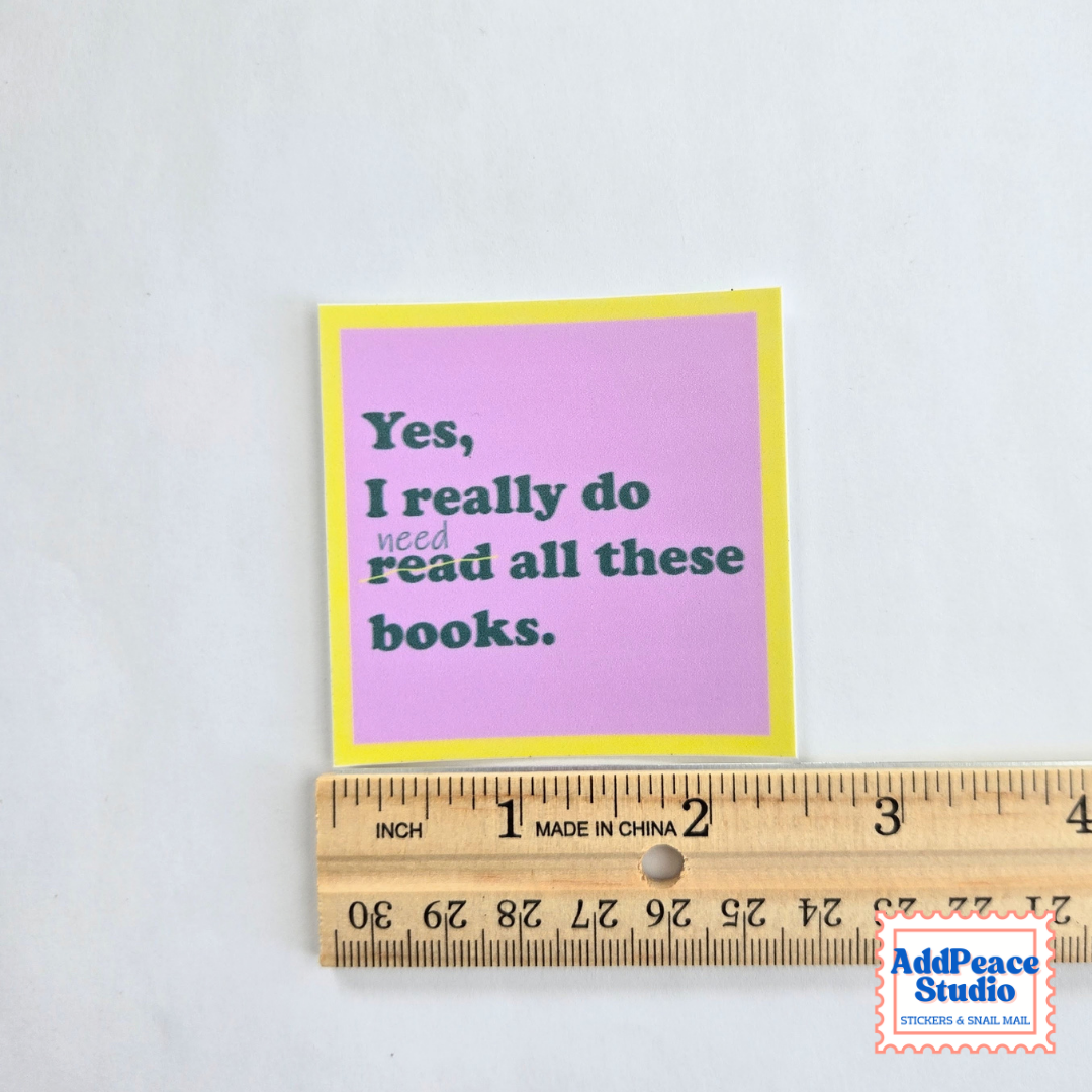 A small square sticker with a pink background, yellow border, and dark green text reading 'Yes, I really do need read all these books.' is shown next to a wooden ruler to indicate size on a white background.