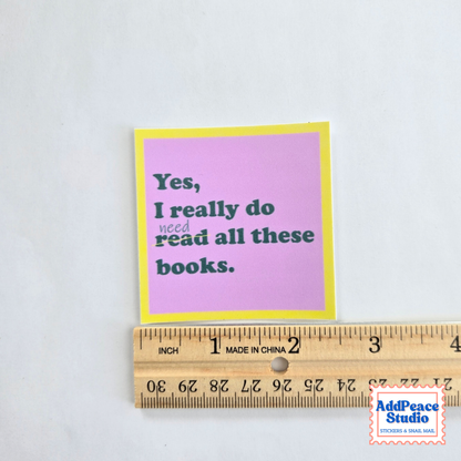 A small square sticker with a pink background, yellow border, and dark green text reading 'Yes, I really do need read all these books.' is shown next to a wooden ruler to indicate size on a white background.