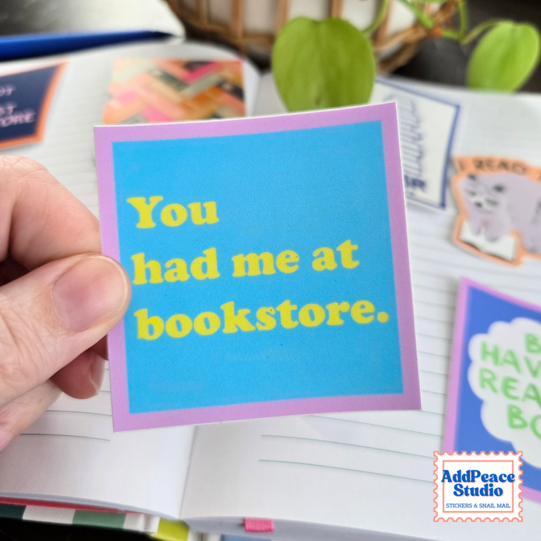 A hand holds a blue square sticker with a purple border and yellow text that says You had me at bookstore. The background is an open notebook with colorful stickers and plants.
