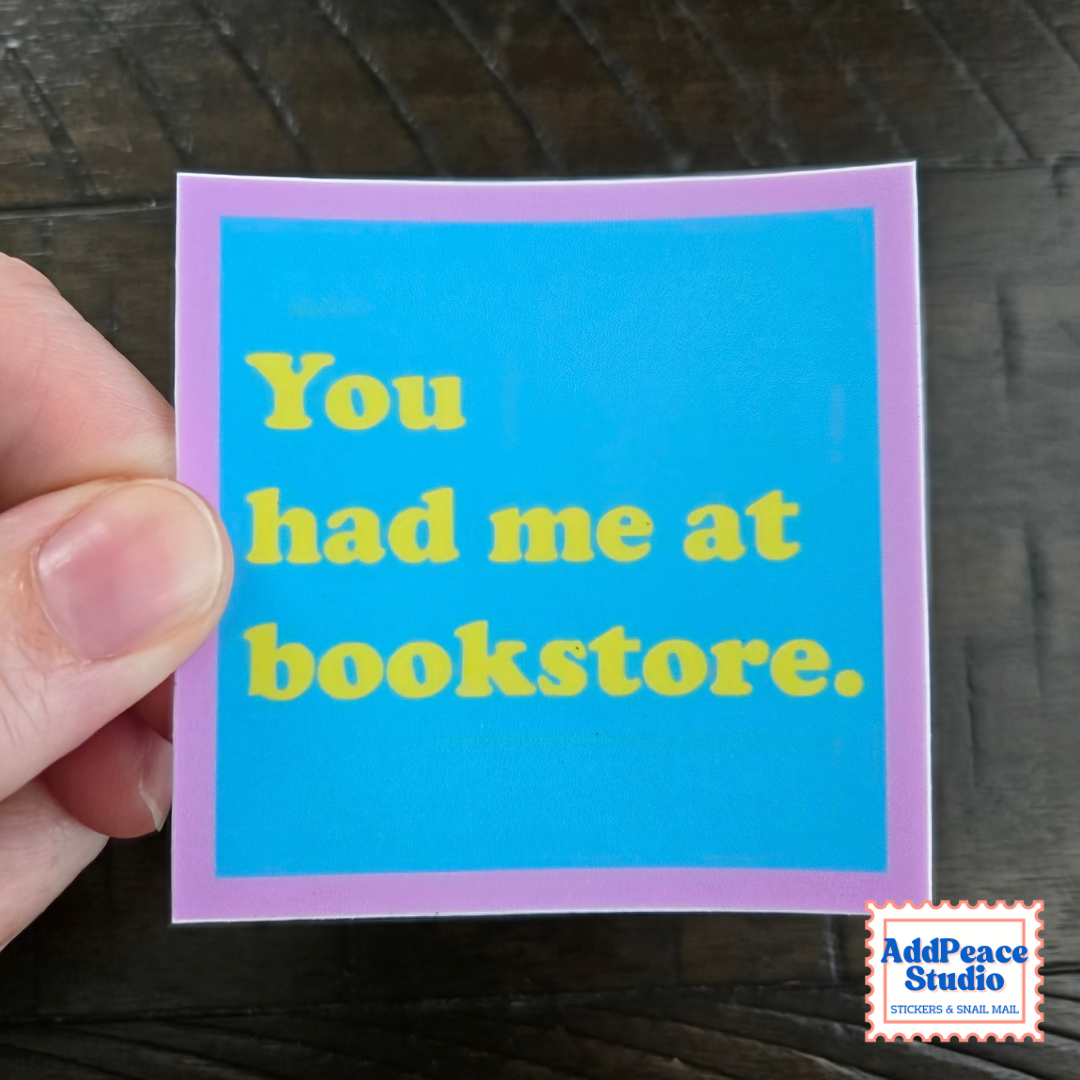 A hand holds a square sticker with a bright blue background, lavender border, and yellow text that reads, 'You had me at bookstore.' A small 'AddPeace Studio' logo appears in the bottom right corner.