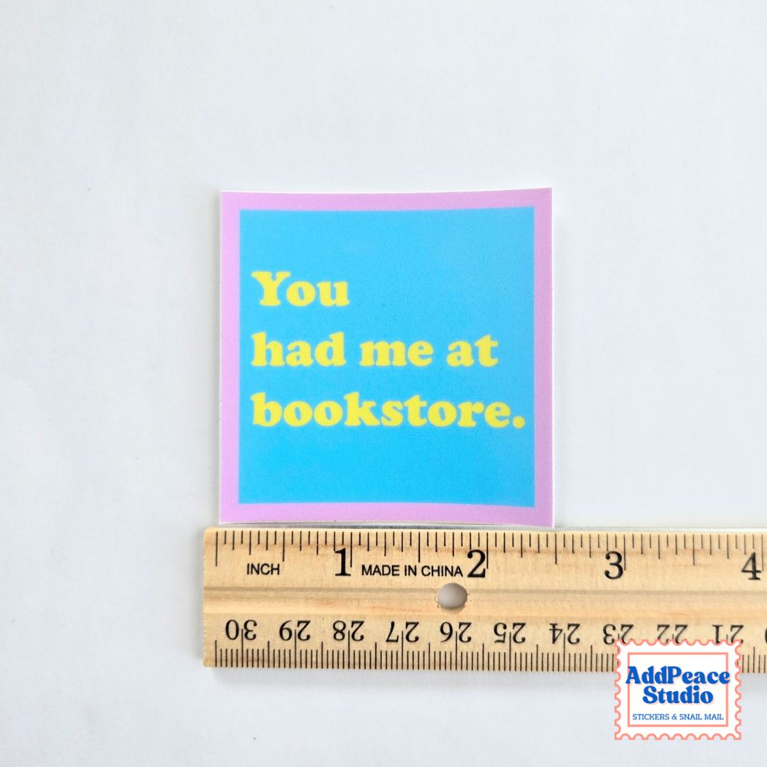 A square printed card or sticker with a bright blue center, pink border, and yellow text reading 'You had me at bookstore.' is shown above a wooden ruler for scale on a white background.