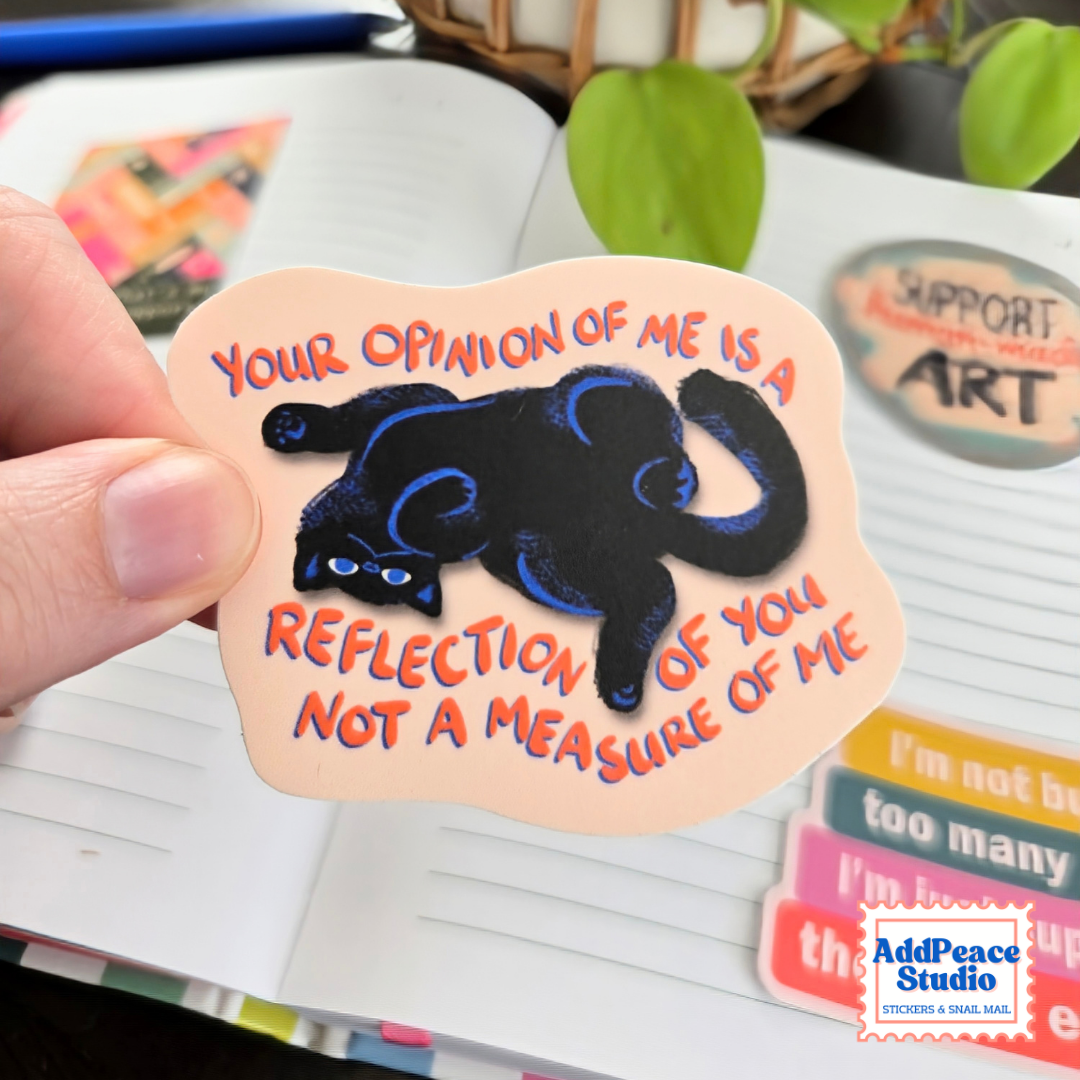A hand holds a die-cut sticker featuring a black cat with blue outlines and orange text that reads, 'YOUR OPINION OF ME IS A REFLECTION OF YOU NOT A MEASURE OF ME.' The sticker is shown in front of a notebook and other colorful stickers, with a visible AddPeaceStudio logo in the bottom right corner.