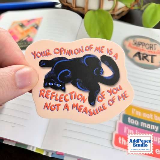 A hand holds a die-cut sticker featuring a black cat with blue outlines and orange text that reads, 'YOUR OPINION OF ME IS A REFLECTION OF YOU NOT A MEASURE OF ME.' The sticker is shown in front of a notebook and other colorful stickers, with a visible AddPeaceStudio logo in the bottom right corner.