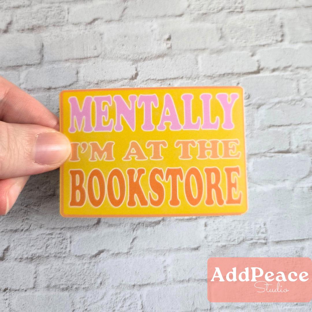 Hand holding a yellow sticker with text 'MENTALLY I'M AT THE BOOKSTORE' against a gray brick wall, with 'AddPeace Studios' logo at the bottom.