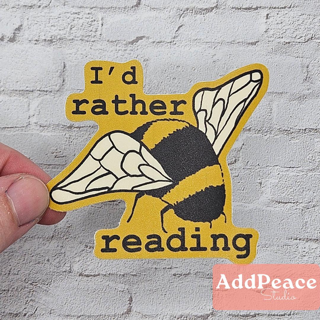 Sticker with a bee design and text 'I'd rather reading' held against a textured wall, with 'AddPeace Studio' branding.