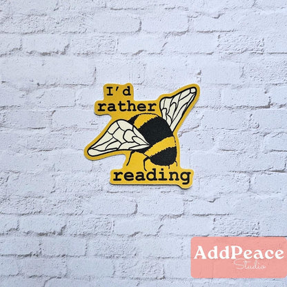 Sticker of a bee with text 'I'd rather reading' on a white brick wall, featuring the brand 'AddPeace Studios'.