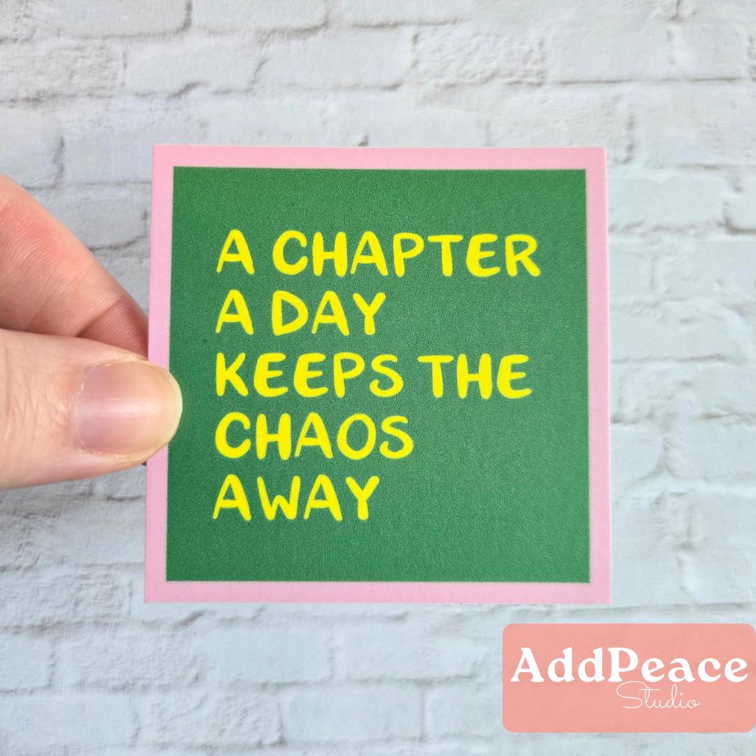 Hand holding a green sticker with yellow text 'A chapter a day keeps the chaos away' against a light gray brick wall.