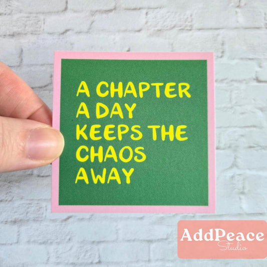Hand holding a green sticker with yellow text 'A chapter a day keeps the chaos away' against a light gray brick wall.