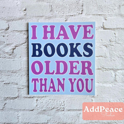 Sticker with 'I HAVE BOOKS OLDER THAN YOU' text on a light blue background, placed against a white brick wall. AddPeace Studios logo at the bottom right corner.