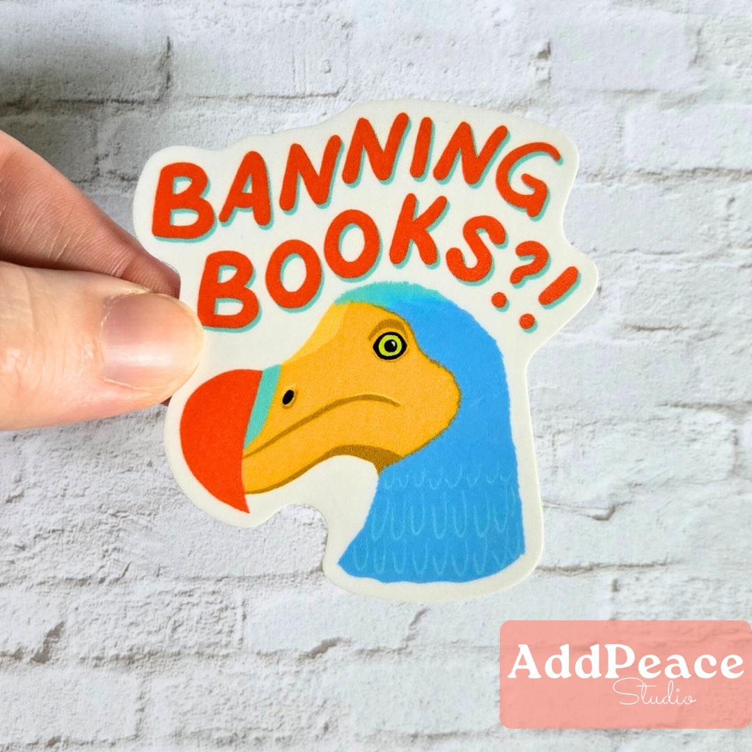 Sticker with a cartoon dodo bird and text 'BANNING BOOKS?!' held by a hand against a light gray brick wall.