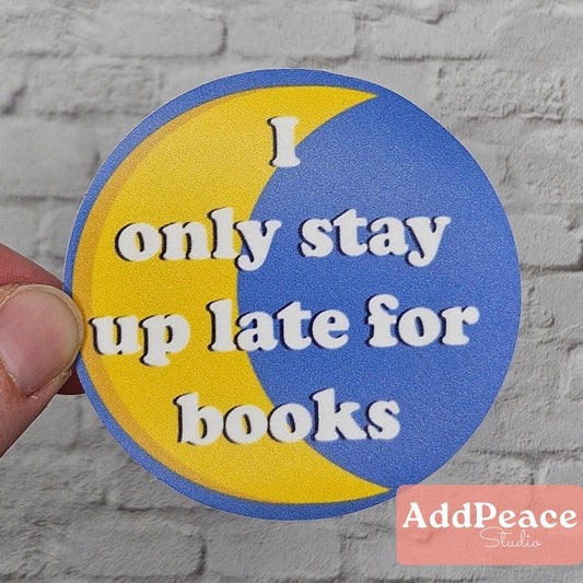 Circular sticker with 'I only stay up late for books' text on a yellow and blue background, held against a brick wall.