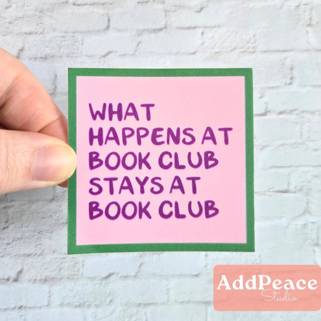 Hand holding a pink sticker with purple text 'What happens at book club stays at book club' against a light gray brick wall.