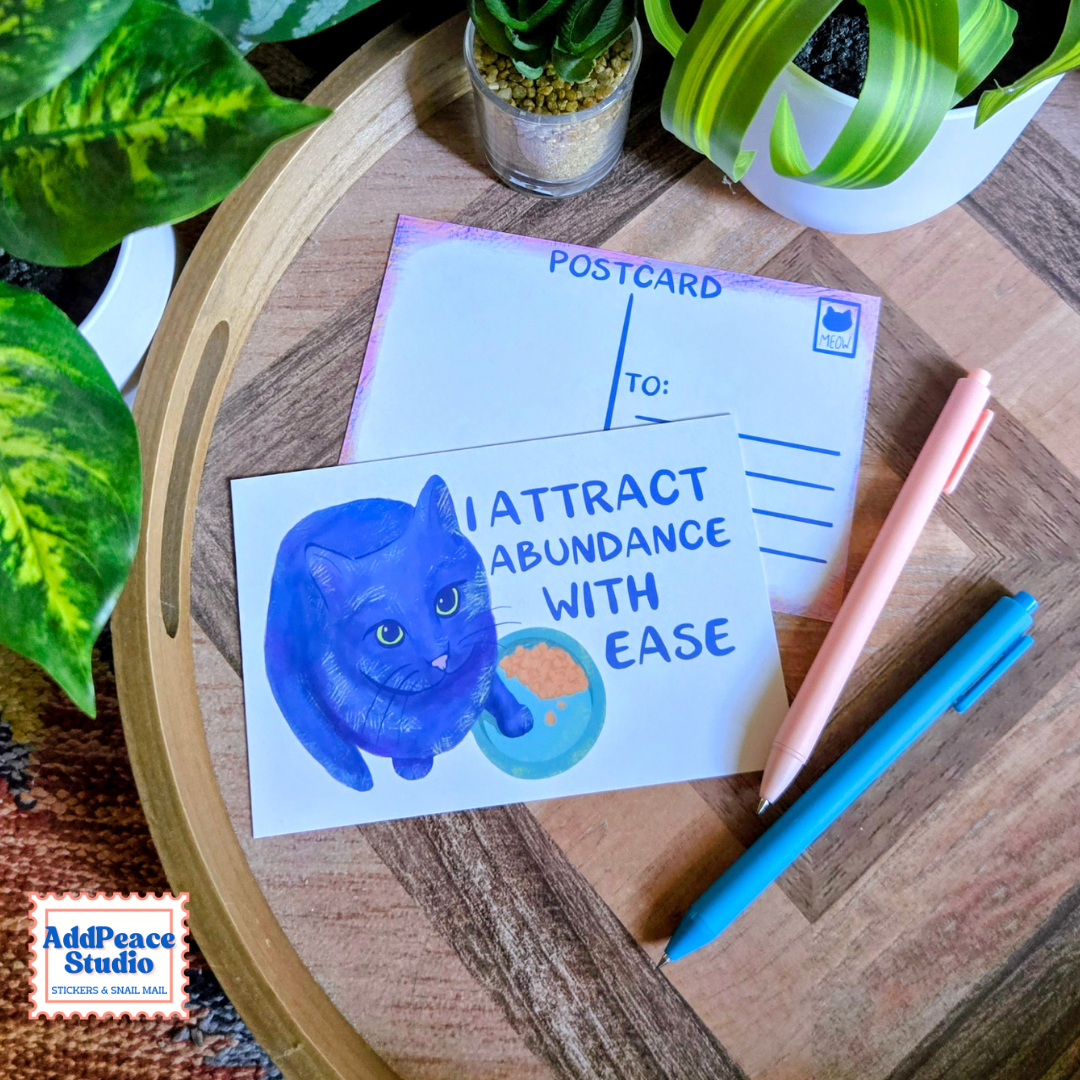 A postcard featuring a blue cat illustration and the text 'I ATTRACT ABUNDANCE WITH EASE' is displayed on a wooden tray with pens and plants.
