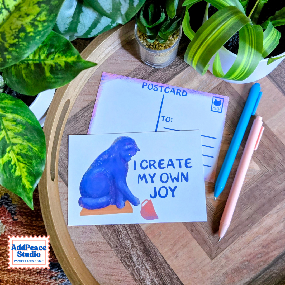 A postcard featuring a blue cat illustration and the text 'I CREATE MY OWN JOY' is displayed on a wooden tray alongside two pens and a small potted succulent. The postcard is branded with the 'AddPeace Studio' logo in the bottom-left corner.