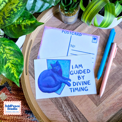 A postcard featuring a blue watercolor cat and the text 'I AM GUIDED BY DIVINE TIMING' is displayed on a wooden tray with pens and potted plants. The postcard is shown alongside the back of another postcard.