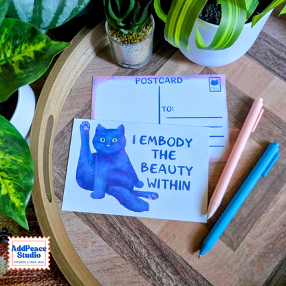 A postcard featuring a blue watercolor cat illustration and the text 'I EMBODY THE BEAUTY WITHIN' is displayed on a wooden tray with pens and potted plants.