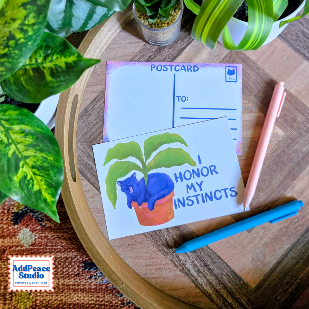 A postcard featuring a blue cat in a pot with the text 'I HONOR MY INSTINCTS' is displayed on a wooden tray alongside a pink pen and a blue pen, with green houseplants in the background.