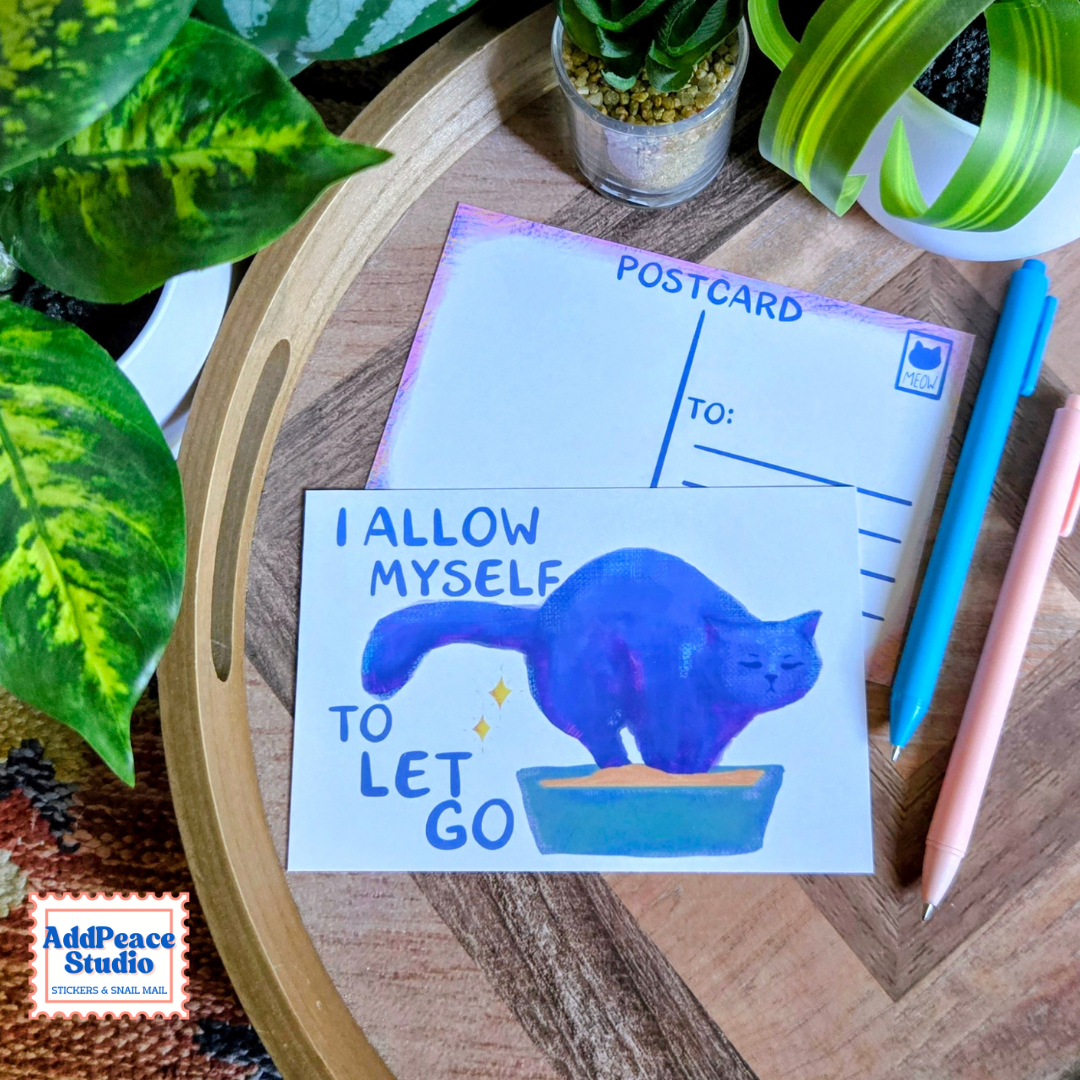 A postcard featuring a blue cat in a litterbox with the text 'I ALLOW MYSELF TO LET GO' is displayed on a wooden tray alongside a postcard writing surface, pens, and green plants.