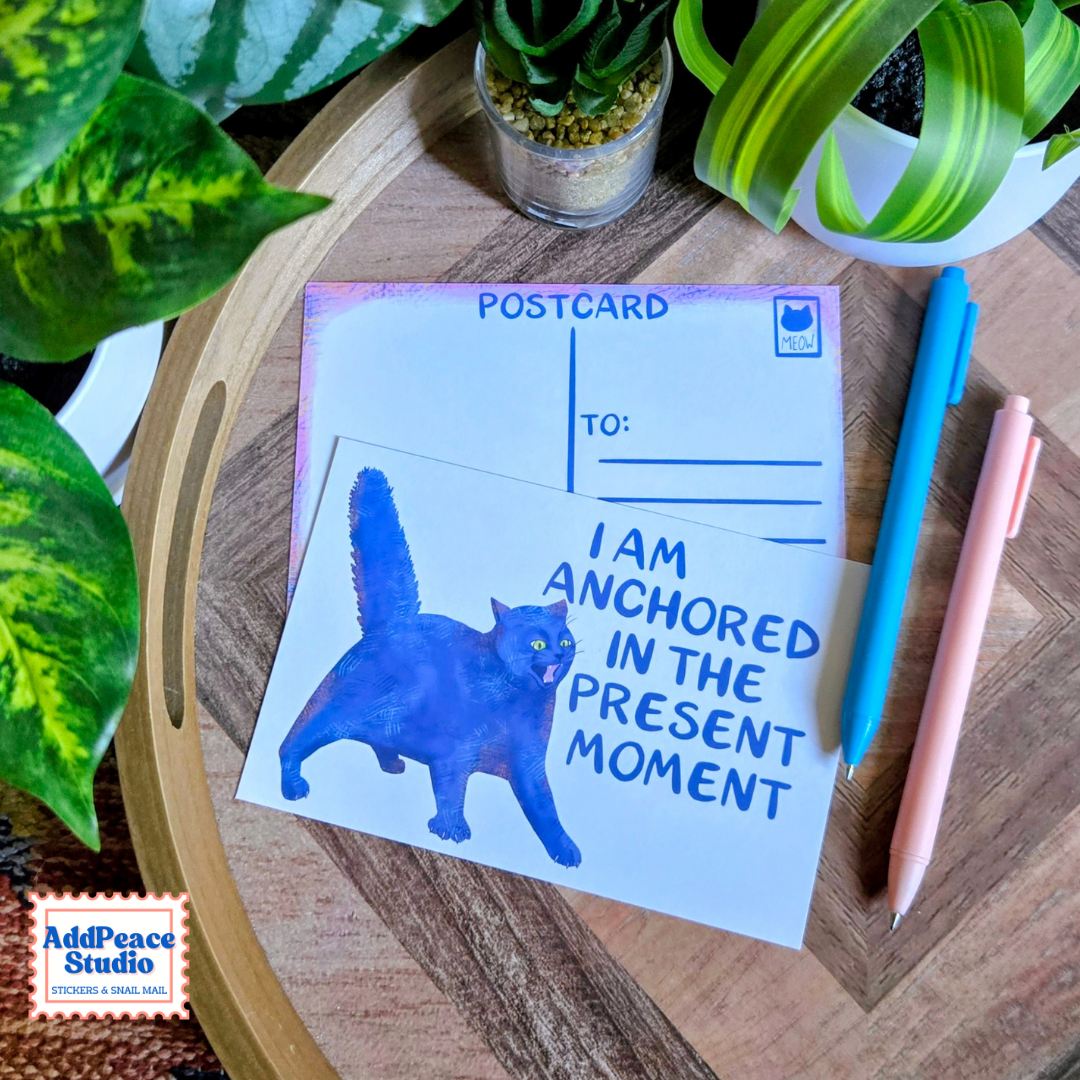 A postcard set styled on a wooden tray with green plants and two pens. The front postcard features a blue watercolor cat and the text 'I AM ANCHORED IN THE PRESENT MOMENT'.