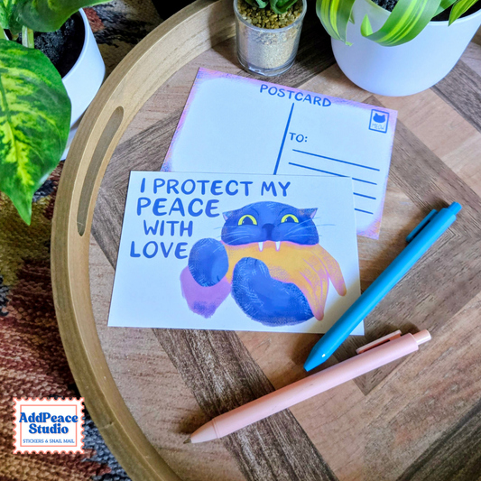 A handmade postcard featuring a colorful painted cat with the text 'I PROTECT MY PEACE WITH LOVE' is displayed on a round wooden tray alongside a blank postcard, two pens, and potted plants. The 'AddPeace Studio' logo is visible in the bottom-left corner.