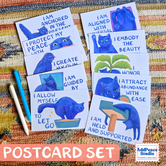A set of printed postcards featuring blue cat illustrations and affirmational phrases, arranged on a patterned fabric background with two colored pencils. A pink banner at the bottom reads 'POSTCARD SET' and the AddPeaceStudio logo appears in the lower right corner.