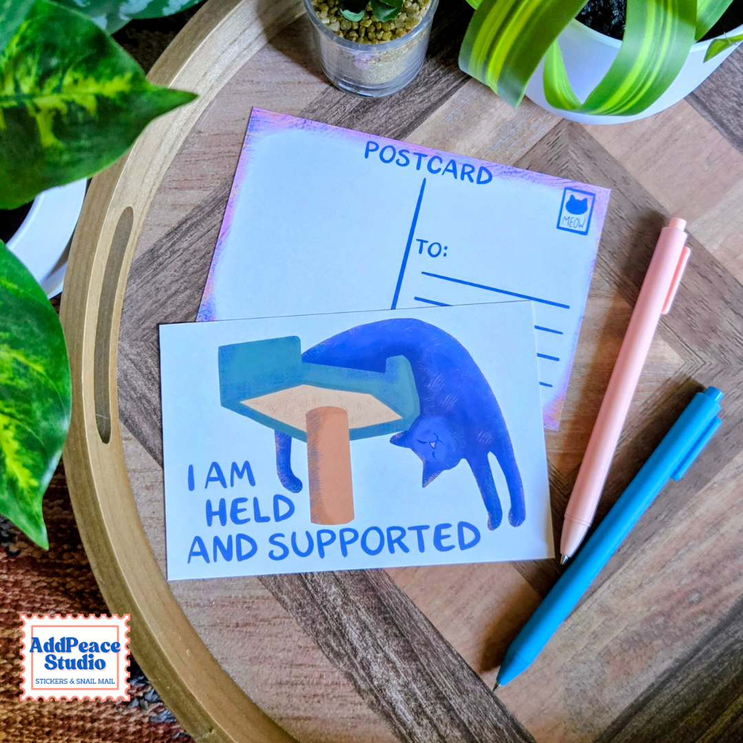 A postcard featuring an illustration of a blue cat leaning against a scratching post with the text 'I AM HELD AND SUPPORTED' is displayed on a wooden tray alongside a postcard back, pens, and green plants.