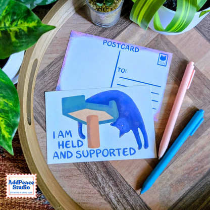A postcard featuring an illustration of a blue cat leaning against a scratching post with the text 'I AM HELD AND SUPPORTED' is displayed on a wooden tray alongside a postcard back, pens, and green plants.