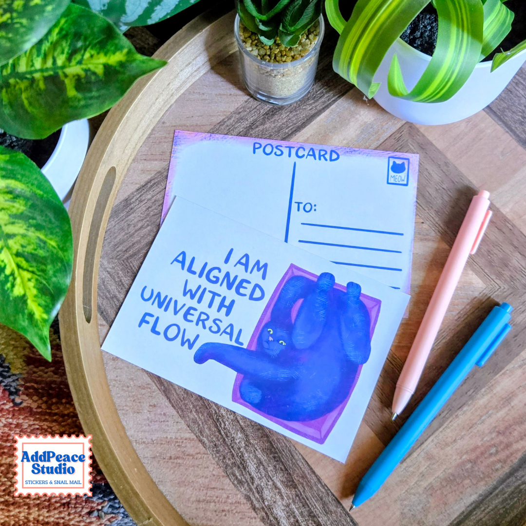 A postcard with a blue cat illustration and the text 'I AM ALIGNED WITH UNIVERSAL FLOW' is displayed on a wooden tray alongside pens and potted plants. The AddPeace Studio logo is visible in the bottom-left corner.