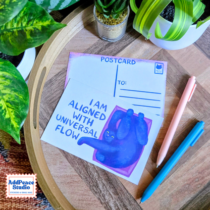 A postcard with a blue cat illustration and the text 'I AM ALIGNED WITH UNIVERSAL FLOW' is displayed on a wooden tray alongside pens and potted plants. The AddPeace Studio logo is visible in the bottom-left corner.
