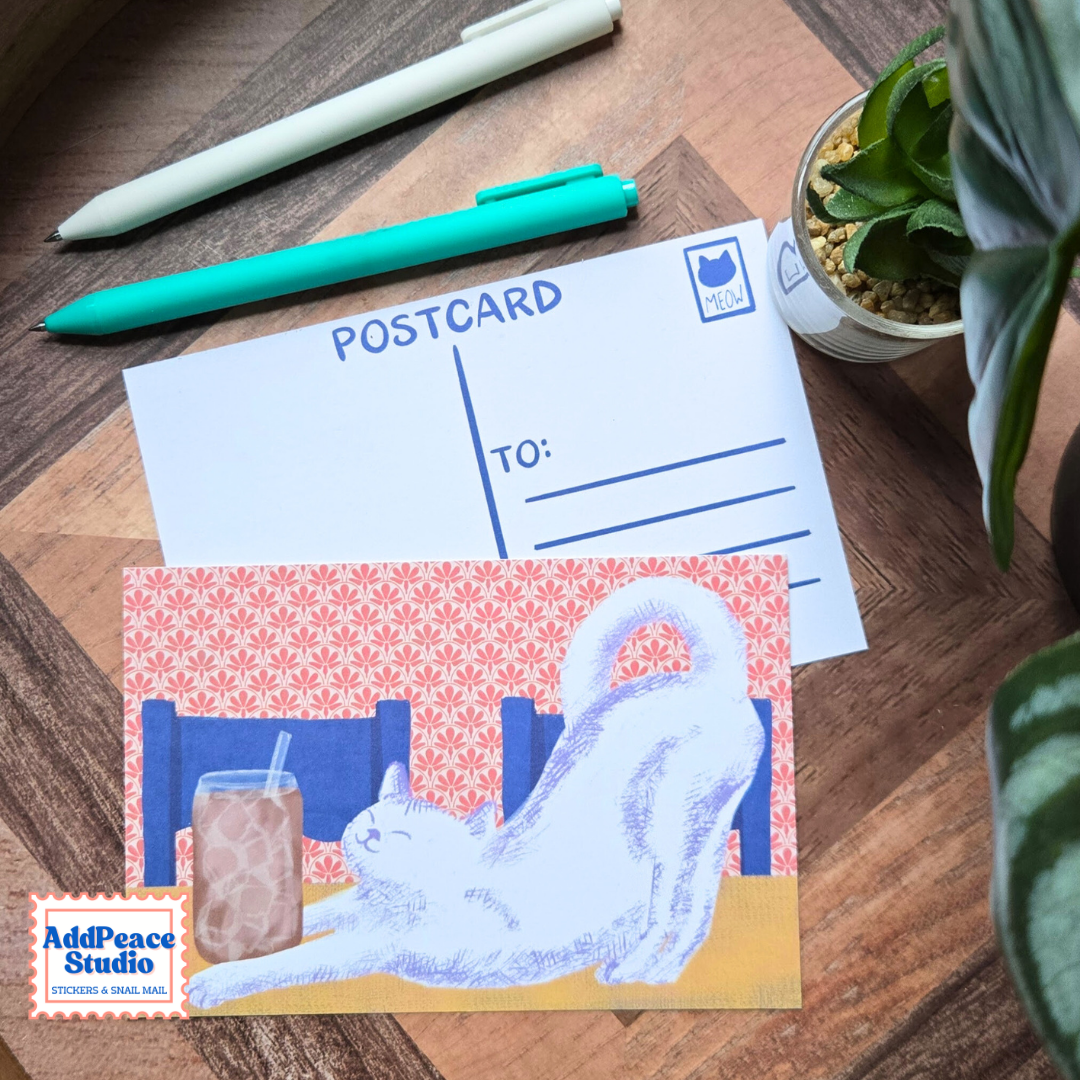 A postcard featuring a white cat stretching in front of an iced coffee, displayed on a wooden desk next to a white postcard template, pens, and a small potted plant. The AddPeace Studio logo is visible in the bottom-left corner.