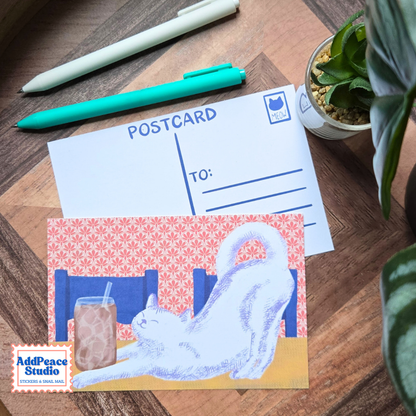 A postcard featuring a white cat stretching in front of an iced coffee, displayed on a wooden desk next to a white postcard template, pens, and a small potted plant. The AddPeace Studio logo is visible in the bottom-left corner.