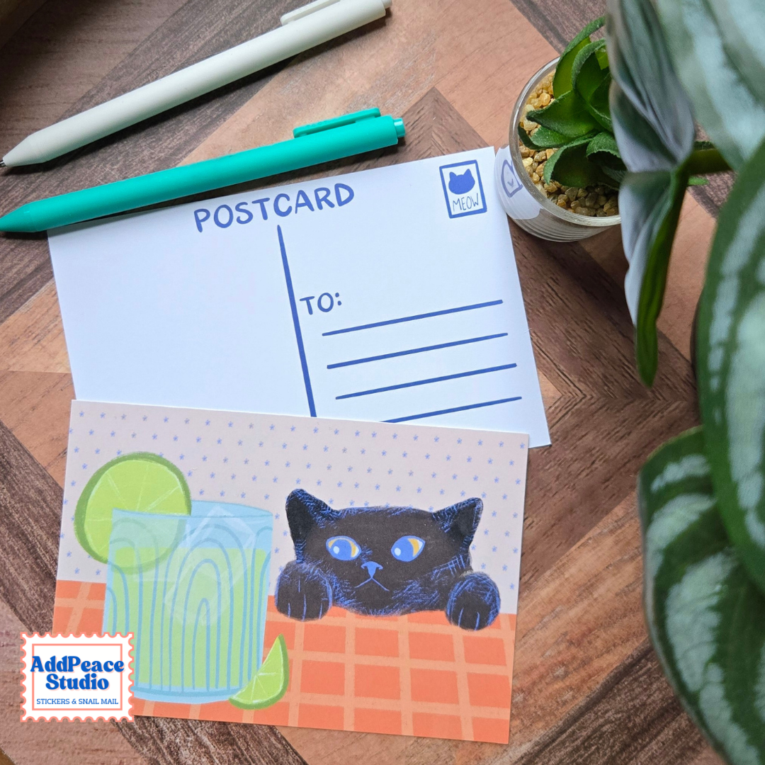 A postcard set displayed on a wooden desk with a white postcard showing blue postcard lines and a colorful illustrated postcard featuring a black cat peeking over an orange table with a glass of limeade. A plant and 2 pens are visible in the scene.