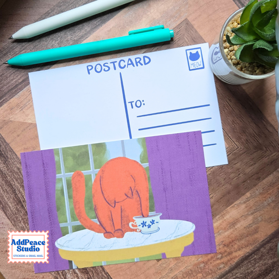 A postcard featuring an illustration of an orange cat sitting at a table with a teacup, layered over a white postcard template with blue lines. The postcards are arranged on a wooden desk alongside pens and a small potted succulent, with the AddPeace Studio logo visible in the bottom left corner.