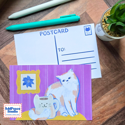 A postcard set displayed on a wooden desk, featuring a white postcard back with blue printing and a colorful illustrated postcard with purple stripes, a white cat, and a small matching cat-shaped mug. Two pens and a small potted succulent are arranged around it.