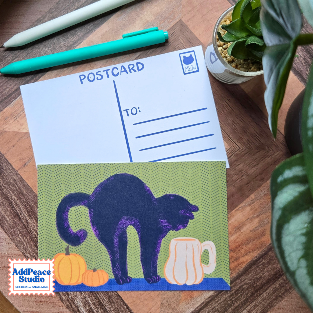 A Halloween-themed postcard featuring a purple cat drinking from a pumpkin-shaped mug with pumpkins beside it, placed on a wooden desk with pens, a plant, and a second postcard showing the writing side.